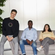 Three people sit on a light gray sofa against a white background. Two men are seated left and center, and a woman sits on the right. They all look toward the camera.