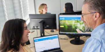 Two people sit at desks with laptops and monitors, while two others talk in the background; business websites are visible on the screens.