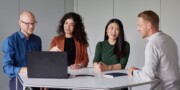 Four people sit at a table having a discussion, with one person pointing at a laptop screen while the others watch and listen.