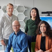 Four people, two standing and two seated, pose and smile in a modern office setting with shelves and a computer screen in the background.