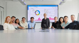 A group of ten people sit and stand around a conference table with laptops, facing the camera; a presentation with graphs is displayed on a large screen behind them.