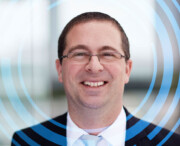 A man wearing glasses and a suit smiles at the camera, with blue concentric circular graphics overlaid on the image.