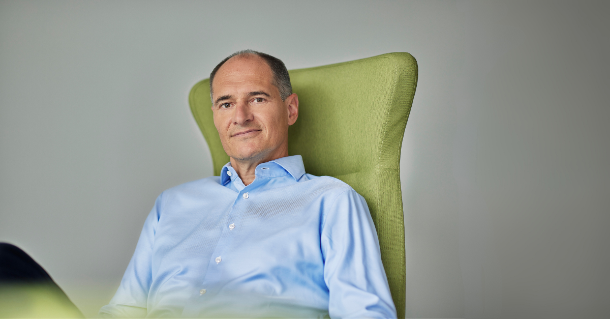 A middle-aged man in a light blue dress shirt sits in a green upholstered chair, looking at the camera with a neutral expression, embodying quiet innovation against a plain background.