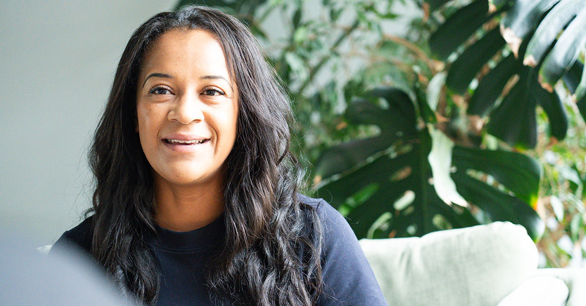A woman with long dark hair smiles while sitting indoors, surrounded by green plants, as she discusses feedback for career growth.