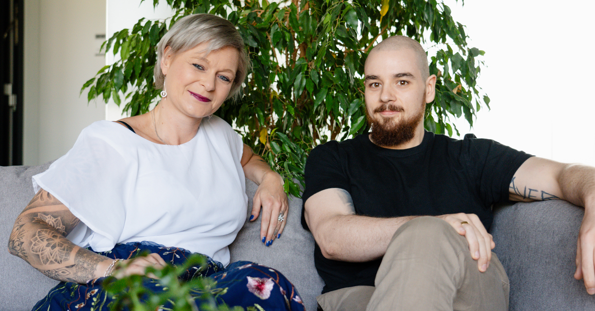Two people sit on a gray couch in front of a green plant—one with short gray hair and tattoos, the other bald with a beard—both looking at the camera, ready to discuss Career Development for IT Leaders.