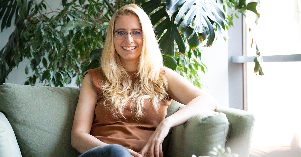 A woman with long blonde hair and glasses sits on a light green couch, smiling confidently, embodying a positive error culture. Large green plants and a window fill the background, creating an inviting atmosphere.