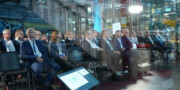 A group of people in business attire sit in rows of chairs at an indoor conference or presentation, with screens and industrial equipment visible in the background.