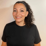 A woman with curly hair in a ponytail, wearing a black t-shirt, stands and smiles in front of a plain white background.