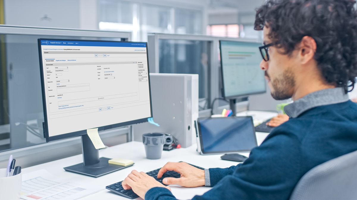 A man with curly hair and glasses works on a desktop computer in a modern office, viewing a form interface on his monitor.