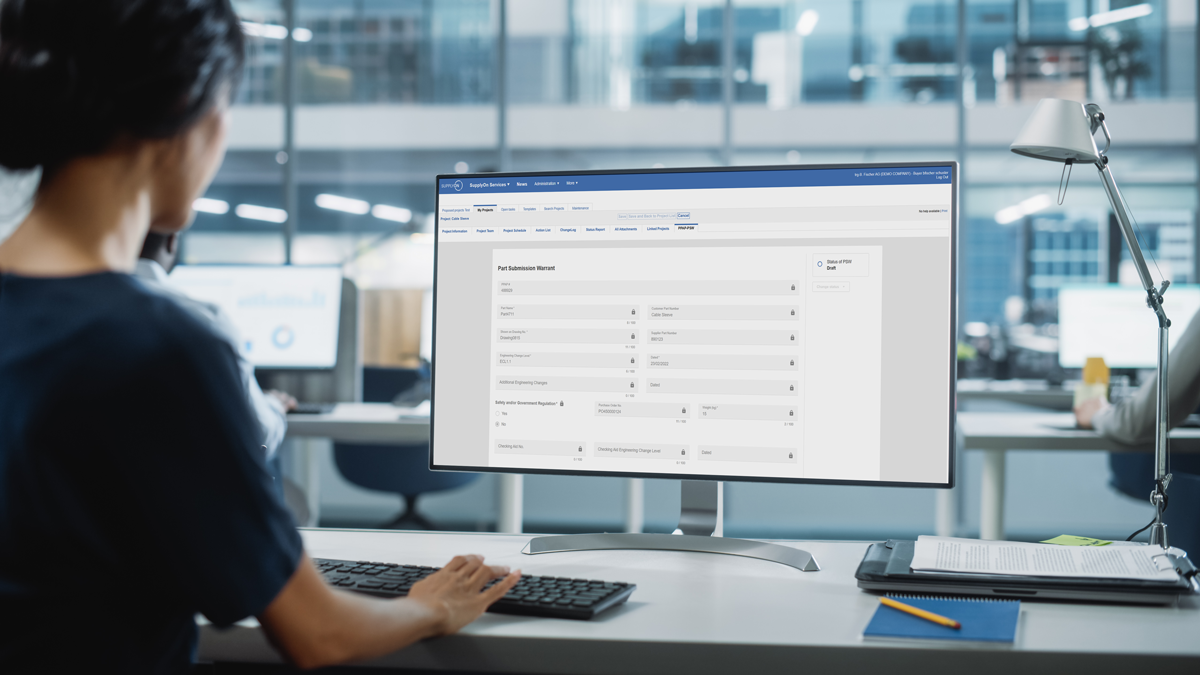 A person sits at a desk in an office, using a computer with a monitor displaying a data entry or reporting form on a business software interface.