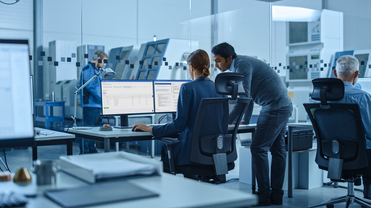 People work at computers in a modern office with glass walls and large monitors, whilst machinery is visible in the background.